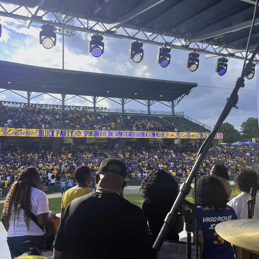 A small band and choir performs on stage during the day with stage lights visible in the background.