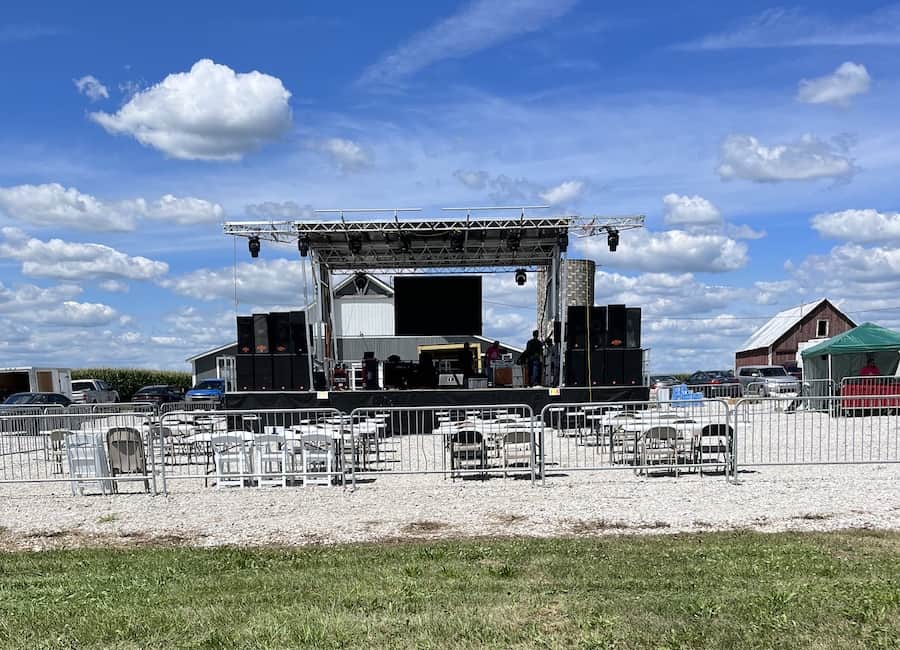 A stage setup in a rural area with a bright blue sky and chairs near a barn.