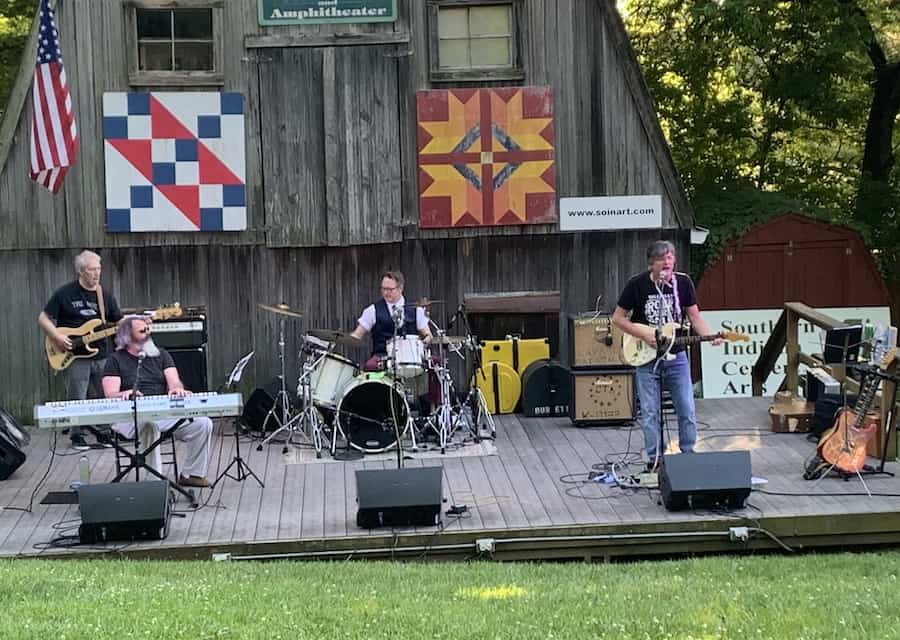 Three artists perform at a rural festival in front of an outdoor pioneer village.