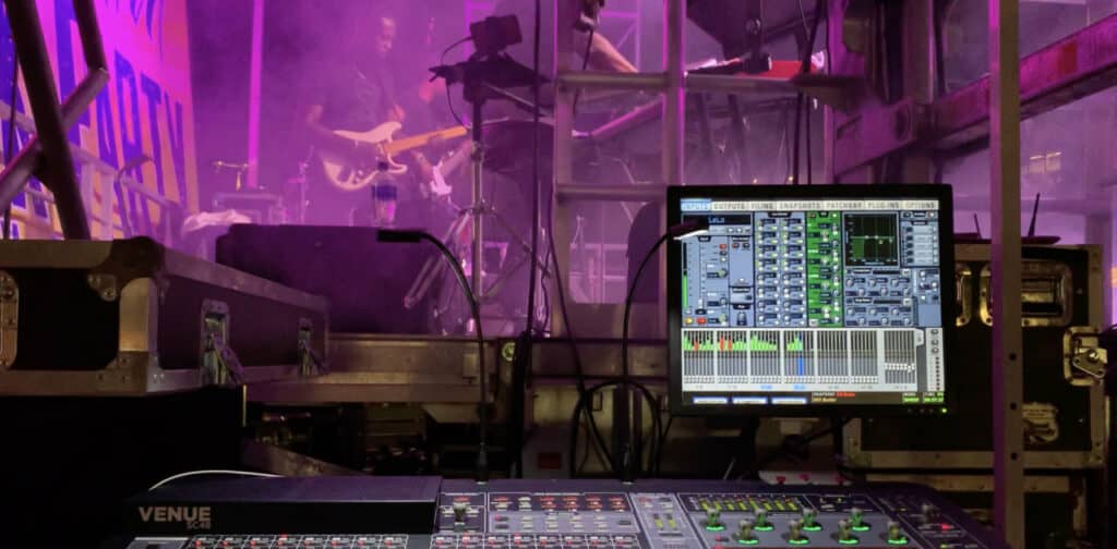 Behind the scenes photo of a guitarist playing with a sound control board and monitor in foreground